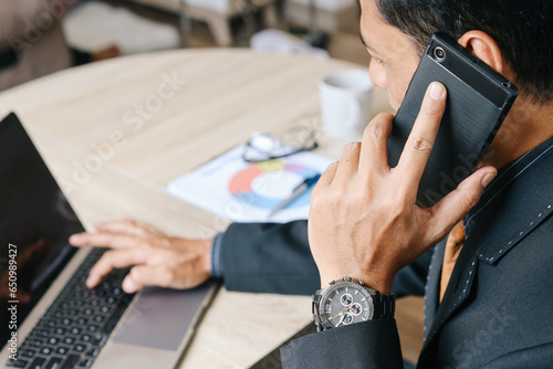 Serious Asian business man sitting on desk using mobile phone while working with laptop at modern office