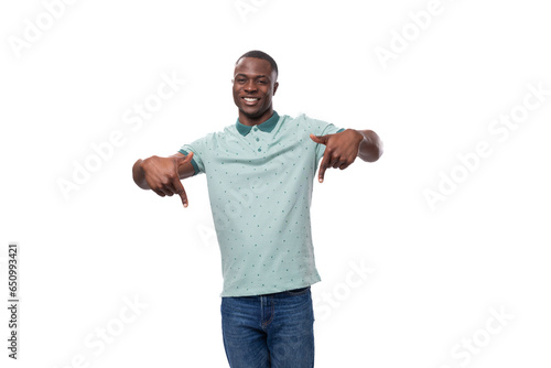 young handsome african guy with short haircut dressed in a mint t-shirt and jeans shows his hands down to the advertising space
