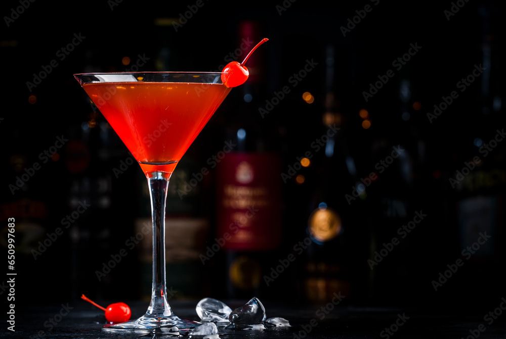 Manhattan cocktail drink with bourbon, red vemuth, bitter, ice and cherry in glass, dark bar counter background, bar tools and bottles