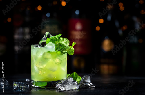 Green Negroni cocktail drink with dry gin, aperitif, gentian liqueur, fresh mint and ice. Black bar counter background, bar tools, bottles