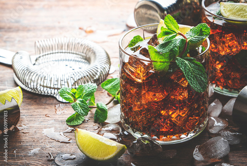 Rum cola cocktail drink with strong alcohol, mint, ice and lime in glass. Wooden background, hard light