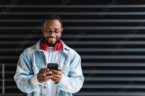 Happy smiling cool gen z young African American ethnic stylish hipster guy model standing at city urban wall using cell phone mobile device, looking at smartphone, holding cellphone.