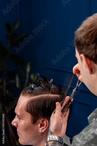 Focused Caucasian male hairdresser cuts a Caucasian guy's hair in a beauty salon. hairdresser at work, blue background, haircut with a typewriter and scissors. 