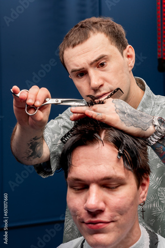 Focused Caucasian male hairdresser cuts a Caucasian guy's hair in a beauty salon. hairdresser at work, blue background, haircut with a typewriter and scissors. 