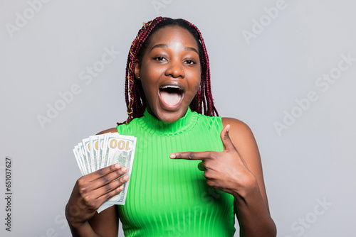 Delighted African American woman with colourful braids smiling holding cash dollars bunch of money in hands celebrating pointing with finger her first salary good payment. photo
