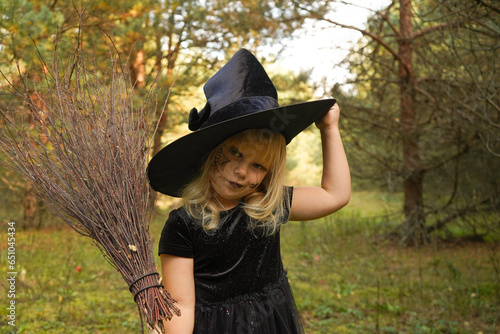 A girl in a witch costume stands with a broom, in an autumn forest. Halloween mysterious lady in woods