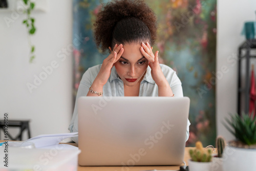 Frustrated Middle Eastern dressmaker with pen and planner leaning on arm while working with laptop in workshop against rack of clothes