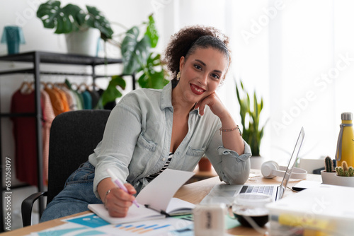 Portrait of optimistic Middle Eastern female seamstress with computer looking at camera and smiling while writing in organizer and working in showroom photo