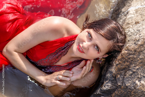 A beautiful elegant lady in a long red dress is swimming in the water near the stones, she is happy.