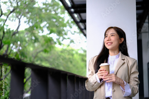 Satisfied millennial businesswoman with paper cup standing outside office building and looking away