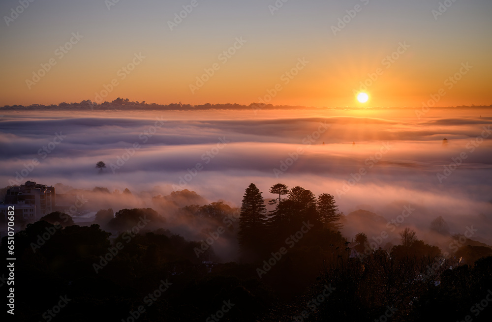 Obraz premium Auckland city above a sea of fog at sunrise. Sun starbursts shining through the clouds. View from the Mt Eden summit. Auckland.
