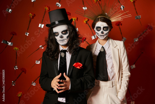 dia de los muertos party, couple in scary makeup looking at camera in red studio with carnations
