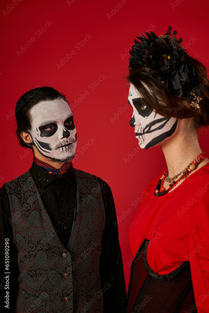 spooky man in sugar skull makeup looking at woman in black wreath, dia de los muertos couple on red spooky man in sugar skull makeup looking at woman in black wreath, dia de los muertos couple on red