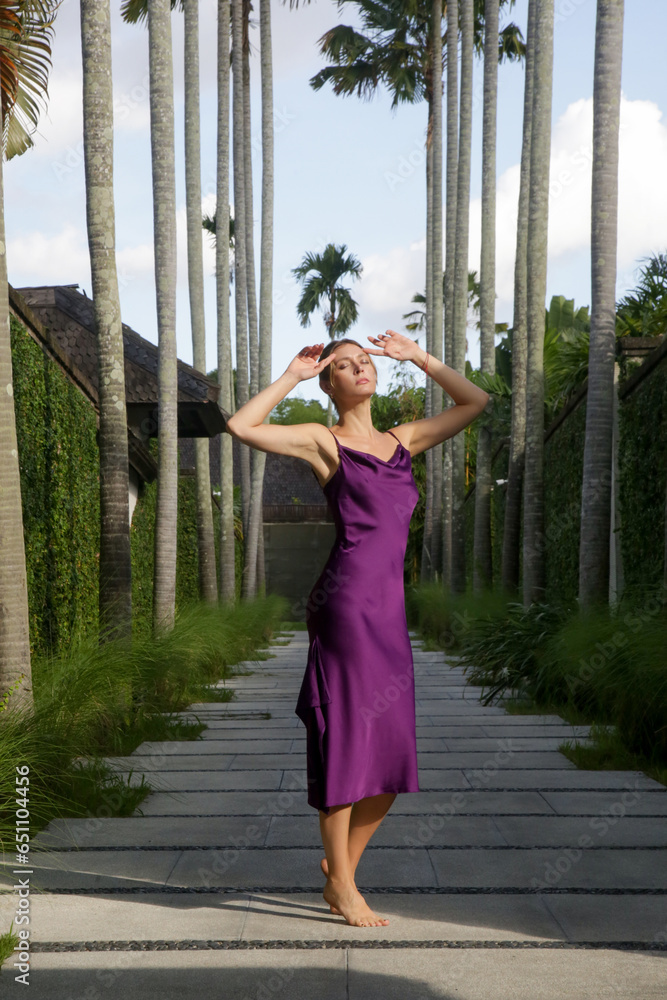Outdoor portrait of stylish fashionable woman in purple silk dress in tropical resort. Natural light. Stylish, elegant summertime outfit, spring-summer fashion trend, tropical vacation style.