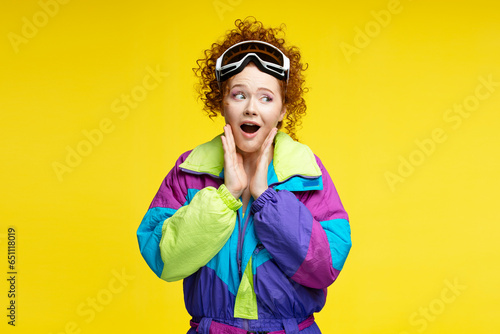 Portrait beautiful excited curly haired woman with open mouth wearing protective ski goggles,  winter overalls, looking away isolated on yellow background photo