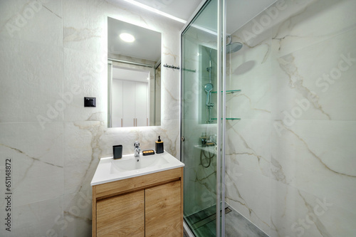 Modern tiled bathroom in white and grey cool colors. It has shower cabin with glass partition, rain shower, wooden stand for white sink and mirror on the wall. Selective focus