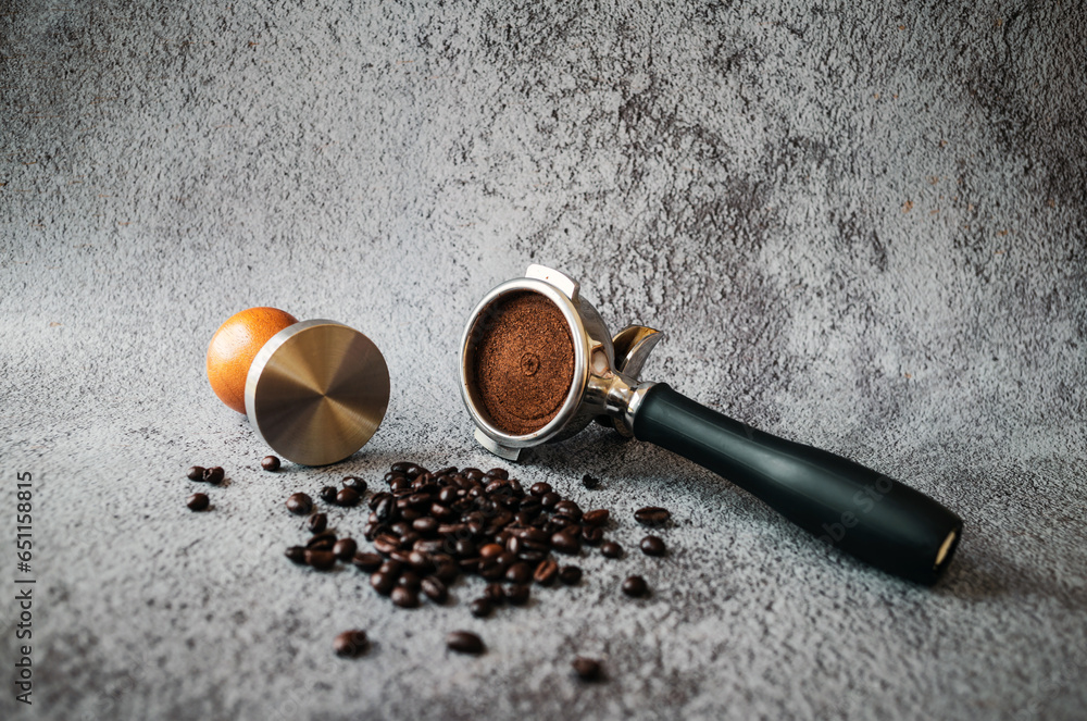 Equipment in a coffee shop of barista coffee tool portafilter with tamper and dark roasted coffee beans on gray background