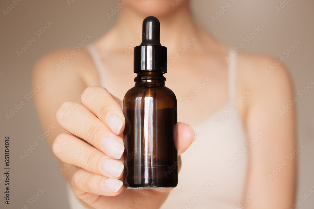 Naklejka premium a woman holds in her hand in front of her a dark glass bottle with a dropper lid on a beige background. Skin care. The concept of beauty. Product advertising