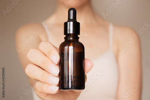 a woman holds in her hand in front of her a dark glass bottle with a dropper lid on a beige background. Skin care. The concept of beauty. Product advertising photo