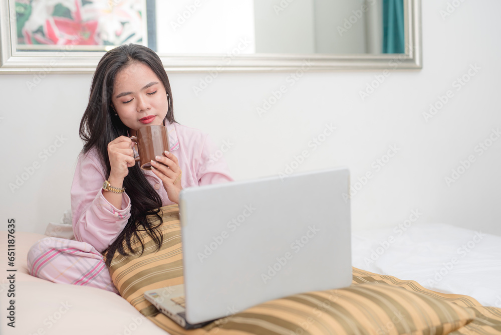 A cute young asian woman having coffee to perk up while working on the laptop on the bed in the morning. Still wearing her pajamas ready to sleep. A cute young asian woman having coffee to perk up while working on the laptop on the bed in the morning. Still wearing her pajamas ready to sleep.