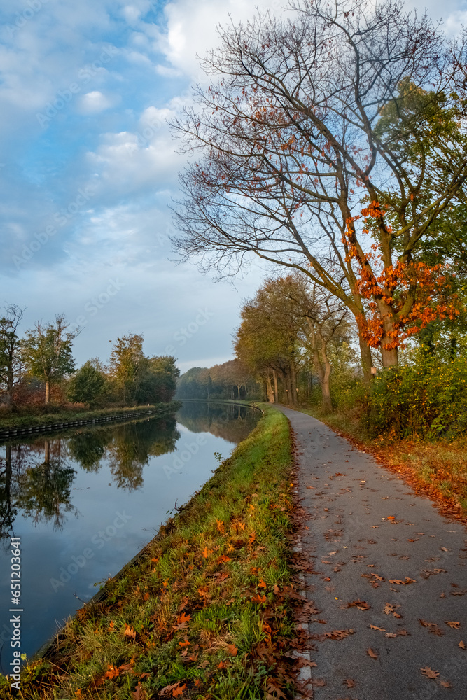 Experience the serene beauty of autumn as you gaze upon this peaceful canal scene. The trees, in various stages of shedding their leaves, create a vibrant carpet of foliage along the water's edge. The Experience the serene beauty of autumn as you gaze upon this peaceful canal scene. The trees, in various stages of shedding their leaves, create a vibrant carpet of foliage along the water's edge. The