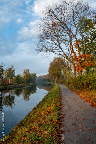 Experience the serene beauty of autumn as you gaze upon this peaceful canal scene. The trees, in various stages of shedding their leaves, create a vibrant carpet of foliage along the water's edge. The