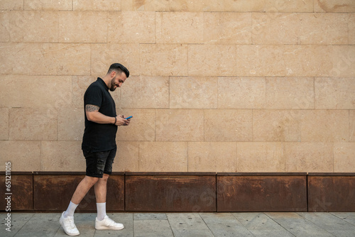 Young smiling man walking along a wall while using his mobile phone.