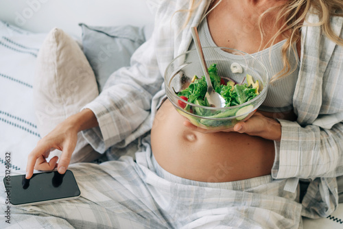 An unrecognizable pregnant woman eats a healthy fresh salad and communicates online on the phone.