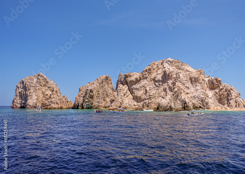 Mexico, Cabo San Lucas - July 16, 2023: Reserva de Lobos Marinos landsend between ocean and Playa de los Amantes, lovers beach under blue sky. Small sightseeing boats in front photo
