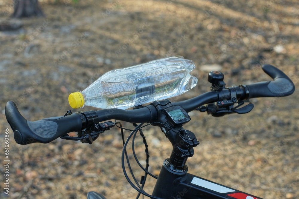 A plastic white bottle of sparkling water closed with a yellow cap lies on the black iron handlebar of a sports modern mountain bike in the forest during the day while resting A plastic white bottle of sparkling water closed with a yellow cap lies on the black iron handlebar of a sports modern mountain bike in the forest during the day while resting