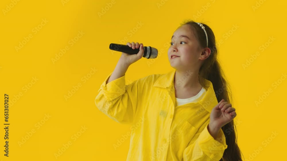 Young asian little girl passionately singing a song to the microphone and smiling on a yellow background. Talented child showing her creative skills Young asian little girl passionately singing a song to the microphone and smiling on a yellow background. Talented child showing her creative skills