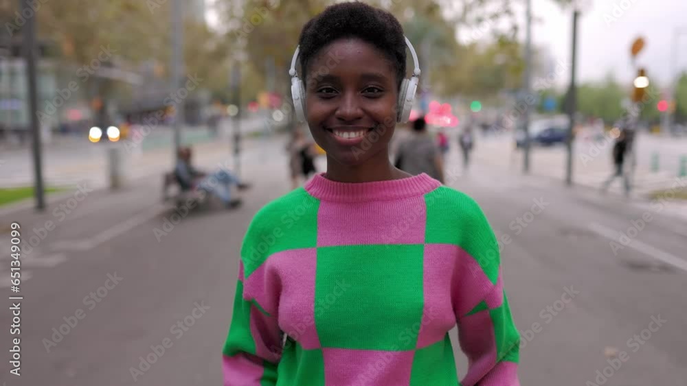 Portrait of cheerful african woman with headphones looking at camera while walking in city street. Urban background, slow motion footage. Portrait of cheerful african woman with headphones looking at camera while walking in city street. Urban background, slow motion footage.