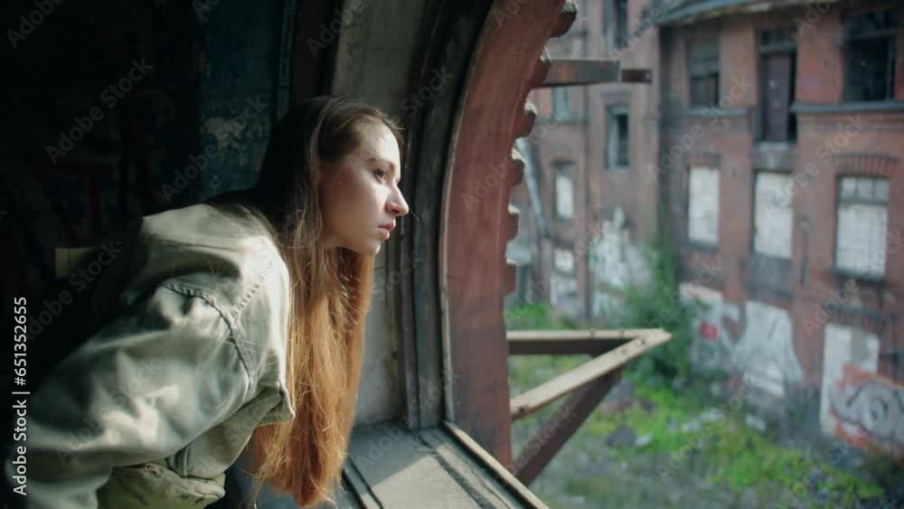 A young caucasian beautiful woman with long hair in green jacket looks thoughtfully through an antique window frame without glass at an old red abondoned destroyed building at sunny day A young caucasian beautiful woman with long hair in green jacket looks thoughtfully through an antique window frame without glass at an old red abondoned destroyed building at sunny day
