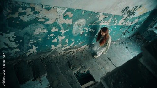 High-angle view of the young caucasian beautiful girl in white jeans and a green jacket climbs the destroyed stairs without steps in an abandoned entrance with blue walls in an old building