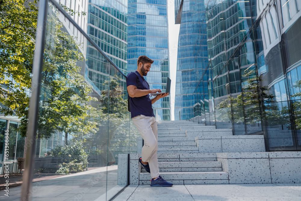 Back view of smiling male manager working on laptop standing on skyscrapers background Back view of smiling male manager working on laptop standing on skyscrapers background