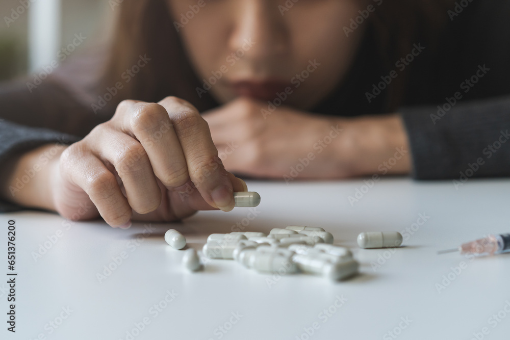 © KMPZZZ - Anti drugs, drug addict asian young woman hand holding pills, medicament with narcotic syringe on table at home, abuse overdose. Sick pain of health, unhealthy people. Suicide depressed or despair.