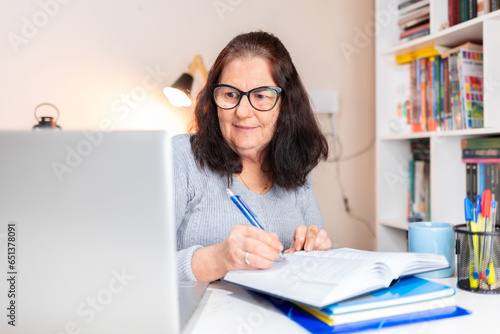 Mature Brazilian woman studying at a home office desk, reading with a laptop beside her, suggesting online learning or internet research. Concept: older individuals returning to education photo