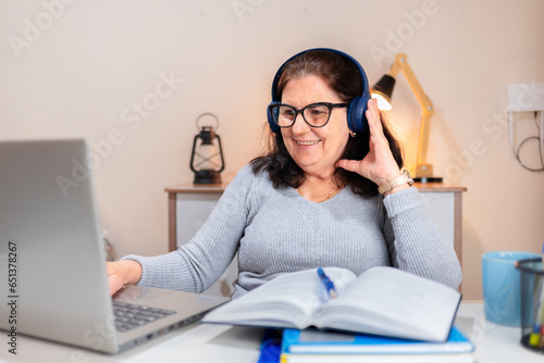 Brazilian mature woman smiling as she uses wireless headphones to study on her laptop at her home office desk. Adults returning to education