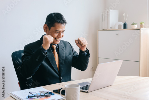 Happy mature Asian businessman clenched fist showing victory gesture while sitting on workdesk in front of laptop at office