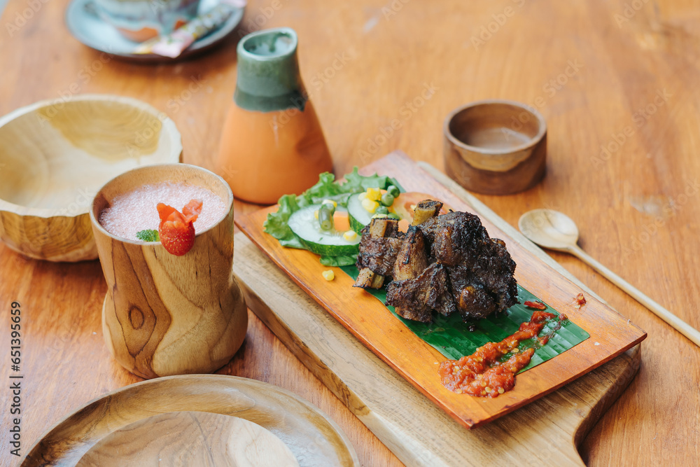 Indonesian food iga bakar or grilled ribs served on wooden plate with vegetable, sambal sauce, and strawberry juice Indonesian food iga bakar or grilled ribs served on wooden plate with vegetable, sambal sauce, and strawberry juice