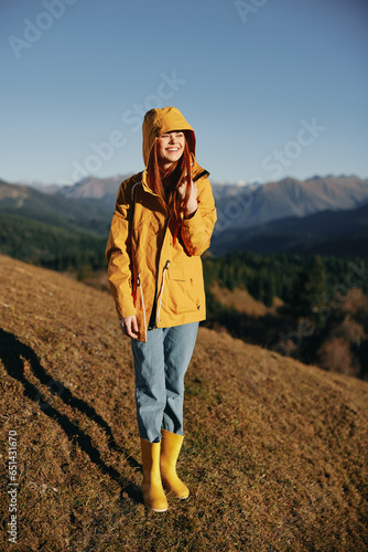 Woman in the autumn smile with teeth full-length walk on the hill and look at the mountains in a yellow raincoat and jeans happy sunset trip on a hike, freedom lifestyle  © SHOTPRIME STUDIO
