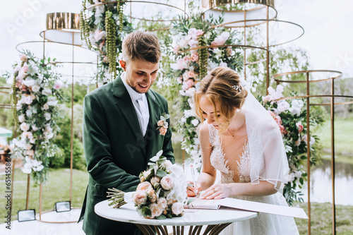 A stylish groom in a green suit and a bride in a white dress sign a marriage registration document against the backdrop of a flower-decorated arch. Wedding photography, portrait.