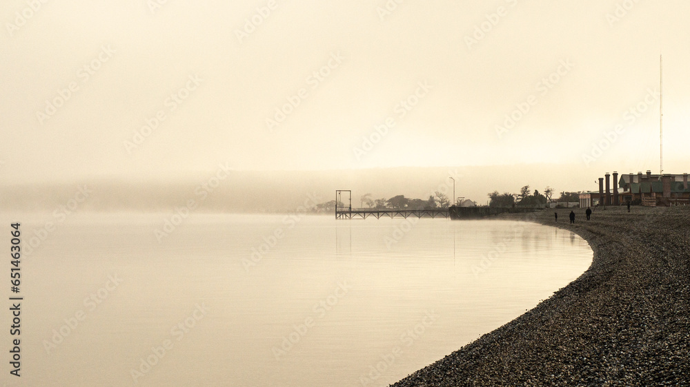 Dock on a lake between fogs with figures of people walking on the shore