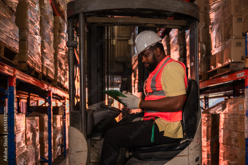 Young black male forklift driver in uniform working in the large logistics warehouse.