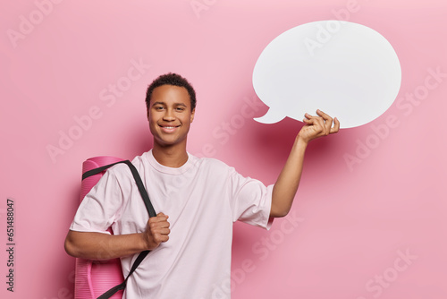 Horizontal shot of handsome curly African man dressed in casual t shrt carries fitness mat holds blank speech bubble for your promotion isolated over pink background. Write your information here photo