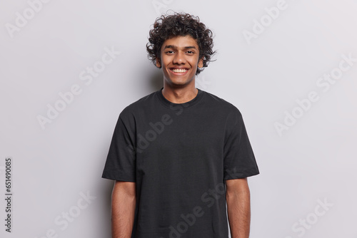 People positive emotions concept. Studio waist up of young happy smiling broadly Hindu man standing in centre isolated on white background wearing black casual t shirt looking straight at camera © Wayhome Studio
