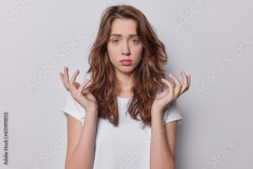 People emotions concept. Indoor waist up of young upset pretty European woman with long brunette hair standing in centre isolated on white background wearing t shirt looking straight at camera