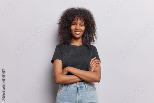 Fényképezés Photo of positive curly haired woman poses with arms crossed smiles gladfully dressed in casual black t shirt and jeans looks directly at camera isolated over white background