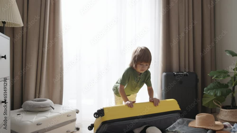 Boy joyfully jumps on packed suitcase. Kid throws plush toy to clothes