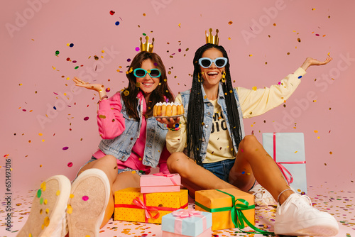 Two joyful young women throwing confetti while holding birthday cake and sitting near gift boxes on pink background